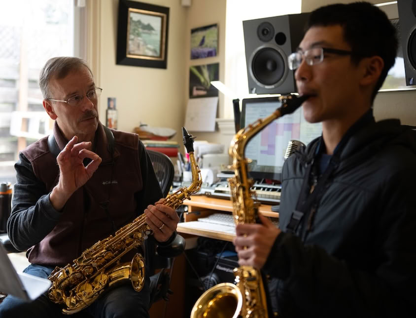 An elderly man instructs a young woman on saxophone in a cozy indoor setting.